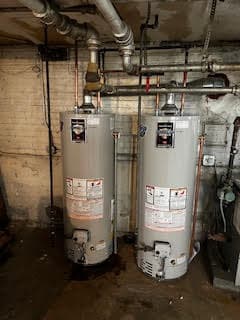 Two gray water heaters installed in a basement, connected with pipes and valves.